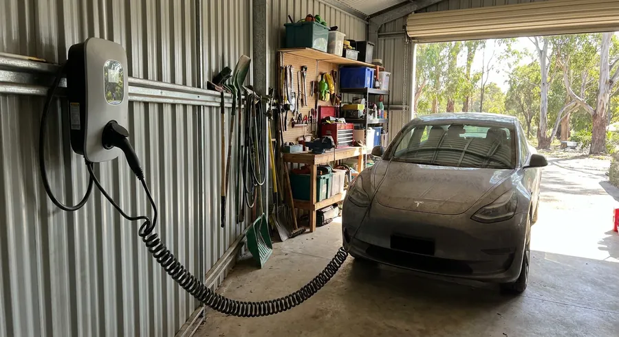 A wall-mounted EV charger in a corrugated iron shed, its coiled cable running to a car parked inside